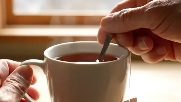 A hand pressing a tea bag against the side of a mug with a spoon, darker color running into the tea